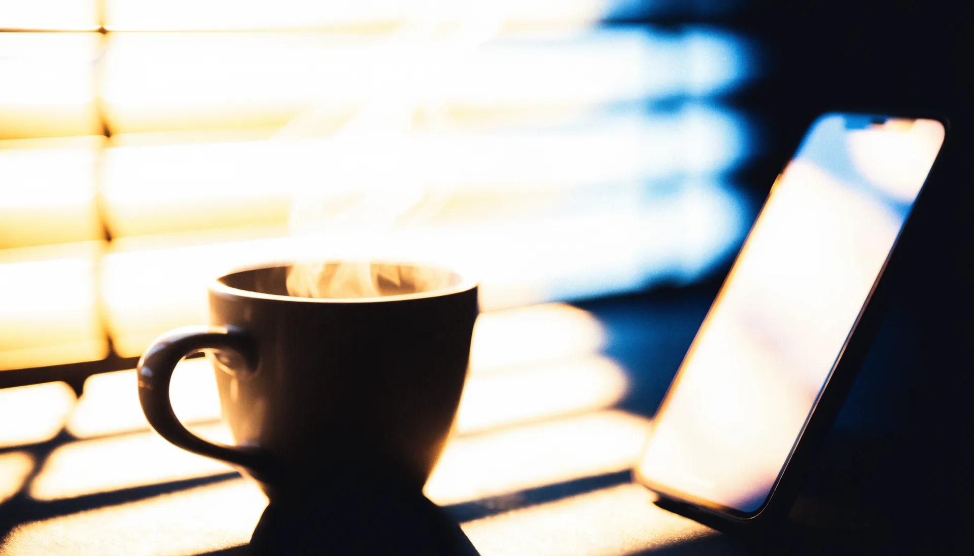 A steaming mug of coffee in gold morning light through the blinds, a phone softly out of focus beside it.