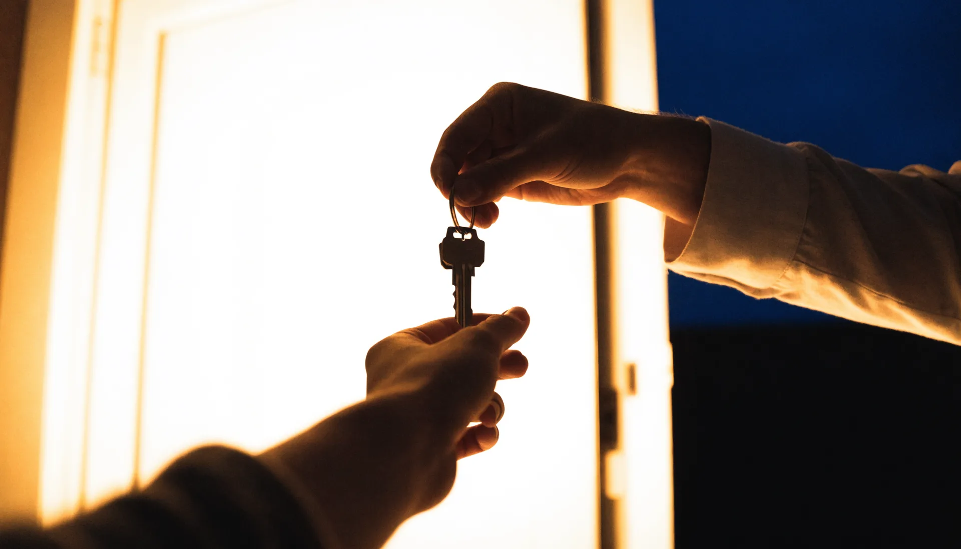 Two hands passing house keys in warm doorway light, the night ink-blue behind them.